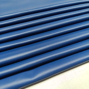 Pode incluir: Close-up de tecido azul escuro dobrado. O material tem uma superfície lisa, ligeiramente texturizada e está disposto em dobras paralelas, criando uma sensação de profundidade e textura. A cor é um azul rico e saturado.