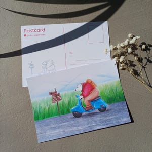 Może przedstawiać: Poczt&oacute;wka z ręcznie rysowaną ilustracją niedźwiedzia na niebieskim skuterze, zmierzającego w stronę znaku z napisem "Mont Blanc". Tył poczt&oacute;wki ma miejsce na adres i znaczek. Ilustracja na tle zielonej trawy i pasma g&oacute;rskiego.