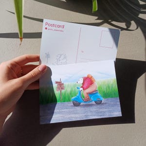 Może przedstawiać: Poczt&oacute;wka z zabawną ilustracją niedźwiedzia polarnego jadącego na niebieskim skuterze w kierunku znaku z napisem "Mont Blanc". Niedźwiedź ma na sobie czerwoną kurtkę i plecak. Poczt&oacute;wka jest oznaczona "Postcard" z nazwiskiem artysty.