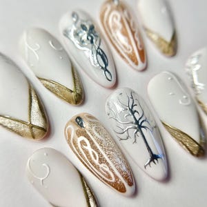 Op de afbeelding: Een set amandelvormige valse nagels. Elke nagel is wit geschilderd met zilveren en gouden accenten. Sommige nagels hebben ingewikkelde zilveren ontwerpen, terwijl andere gouden glitter of gouden punten hebben. De algehele esthetiek is elegant en artistiek.