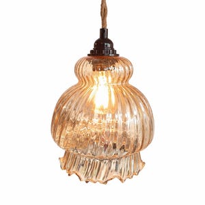 May include: A vintage-style pendant light with a textured, amber-colored glass shade. The shade has a ribbed design and a scalloped edge. It hangs from a brown, twisted cord and a black fixture.