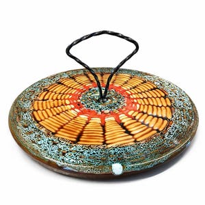 Peut inclure: Un couvercle rond décoratif en verre avec une poignée en métal noir torsadé. Le couvercle présente un motif concentrique dans des tons orange, marron et rouge, avec une bordure mouchetée turquoise et marron. La poignée est une boucle simple et élégante.