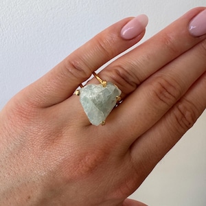 May include: A gold-colored ring featuring a large, irregular-shaped, pale blue-green aquamarine gemstone. The gemstone is held in place by four small prongs. The ring is shown on a hand with pink nail polish.