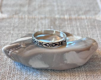 Puede incluir: Un anillo de plata con un patrón detallado en negro y plata. El anillo se muestra sobre una piedra gris y blanca, sobre un fondo de tela neutra. El diseño del anillo presenta elementos geométricos y florales repetidos.
