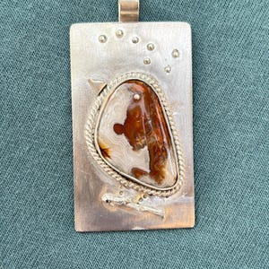 May include: A rectangular silver pendant with a natural stone inlay. The stone is a mix of brown, white, and orange hues, set within a twisted silver rope border. The pendant has a loop at the top for a chain and small silver accents.