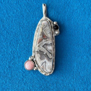Pode incluir: Pingente de prata artesanal com uma pedra central única e estampada. O pingente apresenta uma pedra oblonga grande com redemoinhos cinza, branco e marrom avermelhado, acentuada por uma pedra rosa e preta. O pingente pende de um laço de prata.