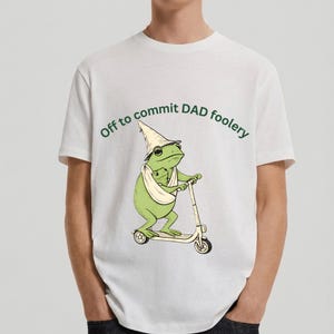 Pode incluir: Camiseta branca com um desenho de um sapo num scooter, usando um chapéu de mago e carregando um bebê sapo. O texto verde acima do sapo diz "Off to commit DAD foolery".