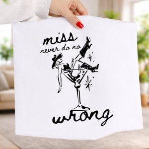 Op de afbeelding: Witte vierkante stof met een zwarte illustratie van een vrouw in een martini-glas, met een hoed en laarzen. De tekst "miss never do no wrong" staat boven en onder de illustratie.