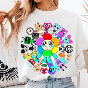 Op de afbeelding: Witte sweatshirt met een kleurrijk cartoonontwerp. De centrale afbeelding is een lachend bloemfiguur met regenboogblaadjes, omringd door andere cartoonfiguren in verschillende vormen en kleuren. Een leuk, speels ontwerp.