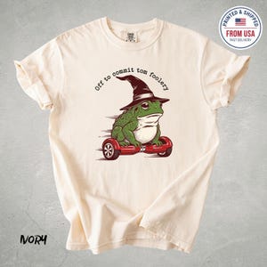 Può includere: T-shirt color avorio con una rana verde con un cappello da strega su un hoverboard rosso. Il testo "Off to commit tom foolery" è sopra la rana. Un logo "Printed & Shipped From USA" è nell'angolo in alto a destra.