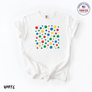 May include: White t-shirt with a square pattern of colorful polka dots in red, green, yellow, and blue. The shirt has a crew neck and short sleeves. The text "WHITE" is at the bottom left.