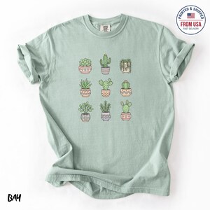 Op de afbeelding: Een lichtgroen T-shirt met negen potcactussen en vetplanten. De planten staan in decoratieve potten met verschillende patronen en kleuren. De tekst "BAY" staat linksonder. Het shirt heeft een relaxte pasvorm. Logo "Printed & Shipped From USA".