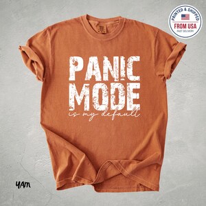 Könnte beinhalten: Gebrannt orangefarbenes T-Shirt mit dem Text "PANIC MODE is my default" in weißen Blockbuchstaben. Das Shirt hat hochgekrempelte Ärmel und einen Rundhalsausschnitt. Ein runder Aufkleber oben rechts lautet "PRINTED & SHIPPED FROM USA FAST DELIVERY".