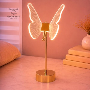 May include: A gold-colored butterfly-shaped LED lamp with illuminated wings. The lamp has a round base and a slender stem. The wings are outlined with a warm white light. The brand name "GLOWNNEST EST. 2024" is visible.