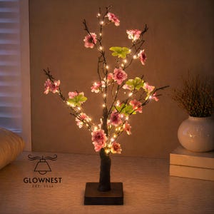 May include: A decorative lighted tree with pink flowers and green leaves. The tree is illuminated with small, warm white lights. The base is a dark brown square, and the trunk is textured. The logo "GLOWNNEST EST 2020" is visible.