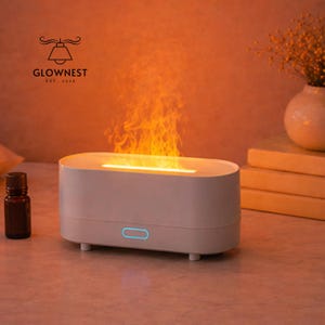 May include: A white, oval aromatherapy diffuser with a flame effect. The diffuser has a blue power button and is next to a small brown essential oil bottle. The logo "GLOWNNEST EST. 2016" is visible in the upper left corner.
