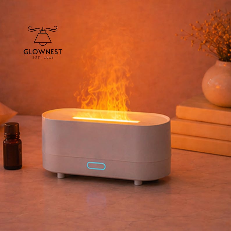 May include: A white, oval aromatherapy diffuser with a flame effect. The diffuser has a blue power button and is next to a small brown essential oil bottle. The logo "GLOWNNEST EST. 2016" is visible in the upper left corner.
