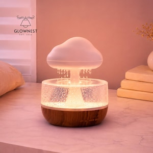 Puede incluir: Una lámpara decorativa en forma de nube con una parte superior blanca y una base transparente texturizada. La base tiene un fondo de vetas de madera. La lámpara está iluminada, creando un brillo cálido. La marca "GLOWNNEST" es visible.