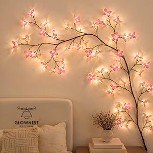 Puede incluir: Una rama decorativa con luces de flores rosas y blancas contra una pared color crema. La rama es de color marrón oscuro y tiene muchas flores pequeñas iluminadas. Las luces crean un ambiente cálido y acogedor. La marca "GLOWNNEST" es visible.