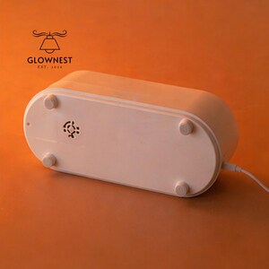 May include: A white, oval-shaped electronic device with rounded edges and four small, circular feet. The bottom of the device features a small vent and a power cord. The logo "GLOWNNEST EST. 2016" is visible in the upper left corner.