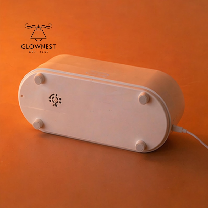 May include: A white, oval-shaped electronic device with rounded edges and four small, circular feet. The bottom of the device features a small vent and a power cord. The logo "GLOWNNEST EST. 2016" is visible in the upper left corner.