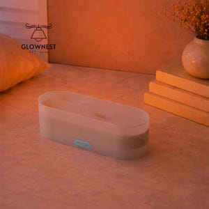 May include: An off-white rectangular essential oil diffuser with a blue power button. The diffuser sits on a light-coloured surface. The GlowNest logo is visible. A small vase with dried flowers is in the background.