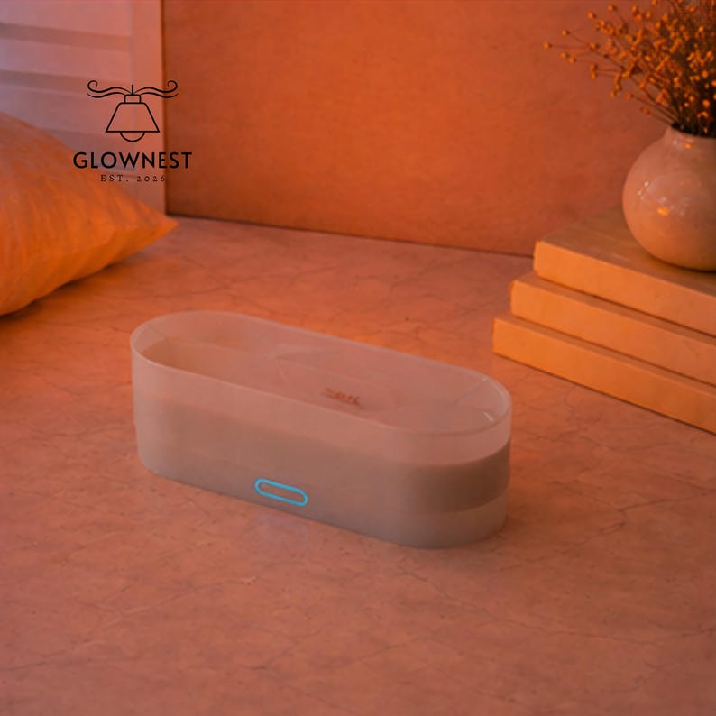 May include: An off-white rectangular essential oil diffuser with a blue power button. The diffuser sits on a light-coloured surface. The GlowNest logo is visible. A small vase with dried flowers is in the background.