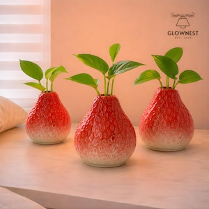 May include: Three red strawberry-shaped ceramic vases with green plants. The vases have a textured surface with white and red gradients. The vases are on a white surface. The GlowNest logo is in the top right corner.