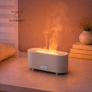 May include: A white oval aromatherapy diffuser with a flame effect. The diffuser has a blue light and is emitting a flame-like vapor. The GlowNest logo is in the upper left corner. A vase of dried flowers and books are in the background.
