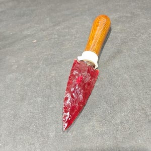 May include: A dagger featuring a translucent, deep red, flaked stone blade with a sharp point. The handle is made of polished, light brown wood with a visible grain, connected to the blade by a white, cylindrical collar piece.