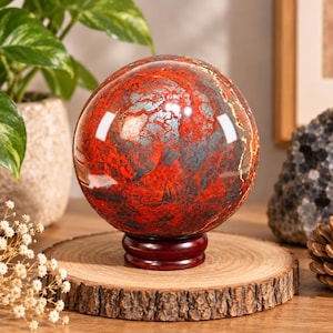 Peut inclure: Une pierre sphérique polie présentant un motif marbré rouge vif et gris foncé avec de subtiles veines métalliques. La sphère repose sur un petit support circulaire en bois brun foncé, affichant une surface lisse et réfléchissante.