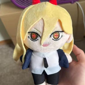 May include: A plush doll of a character with yellow hair, red horns, and detailed facial features. The doll wears a white shirt, black pants, and red shoes. The doll's eyes have a cross design.