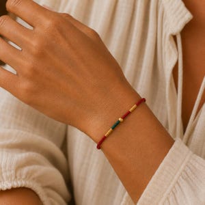Puede incluir: Una pulsera fina con una base de cordón rojo acentuada con dos pequeñas cuentas cilíndricas doradas, una cuenta cilíndrica verde y una pequeña cuenta esférica dorada situada entre los elementos cilíndricos verdes y dorados.