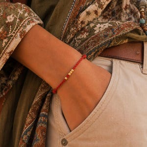 Könnte beinhalten: Ein rotes Schnurarmband mit drei kleinen goldenen Perlen. Das Armband ist an jedem Ende geknotet. Die Person trägt eine khakifarbene Hose und einen gemusterten Schal.