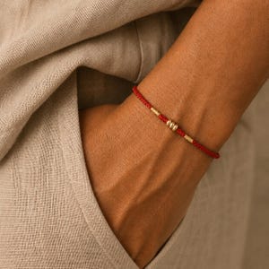 May include: A red string bracelet with three gold beads. The bracelet is worn on a wrist, with a neutral-colored fabric in the background. The bracelet is a simple, elegant piece of jewelry.
