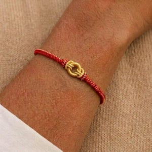 Peut inclure: Bracelet rouge et or avec un motif de nœud. Le bracelet est composé d'un cordon rouge avec des accents dorés et un élément central en forme de nœud doré. Le bracelet est porté au poignet.