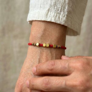 Puede incluir: Pulsera de hilo rojo con cuentas cuadradas doradas y cuentas más pequeñas doradas y plateadas. La pulsera se lleva en la muñeca, con una manga de camisa blanca en el fondo. Un accesorio sencillo y elegante.