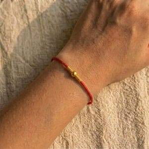 May include: A red string bracelet with a gold bead in the center. The bracelet is worn on a person's wrist. The background is a light beige fabric.