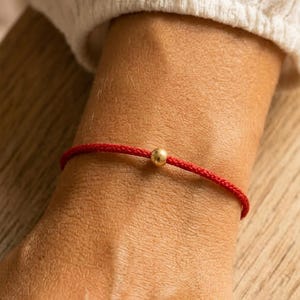 Op de afbeelding: Een rode touw armband met een goudkleurige kraal. De armband is eenvoudig en minimalistisch, met de kraal gecentreerd op het touw. De armband wordt om een pols gedragen, en de achtergrond is wazig.