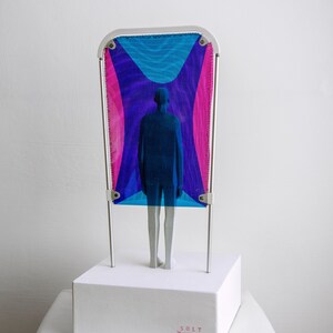 Puede incluir: Una escultura con una base rectangular blanca que tiene el texto SOLT 2025 impreso en rojo. Un marco de metal plateado sostiene una malla con patrones geom&eacute;tricos azules y rosas. Una figura humana azul se encuentra frente a la malla.