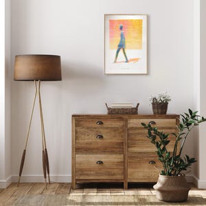 K&ouml;nnte beinhalten: Ein gerahmter Kunstdruck mit einer stilisierten Figur, die vor einem leuchtend gelben und orangen Hintergrund geht, an einer Wand &uuml;ber einer h&ouml;lzernen Kommode mit sechs Schubladen, einer Stehlampe, einem geflochtenen Korb und einer Pflanze.