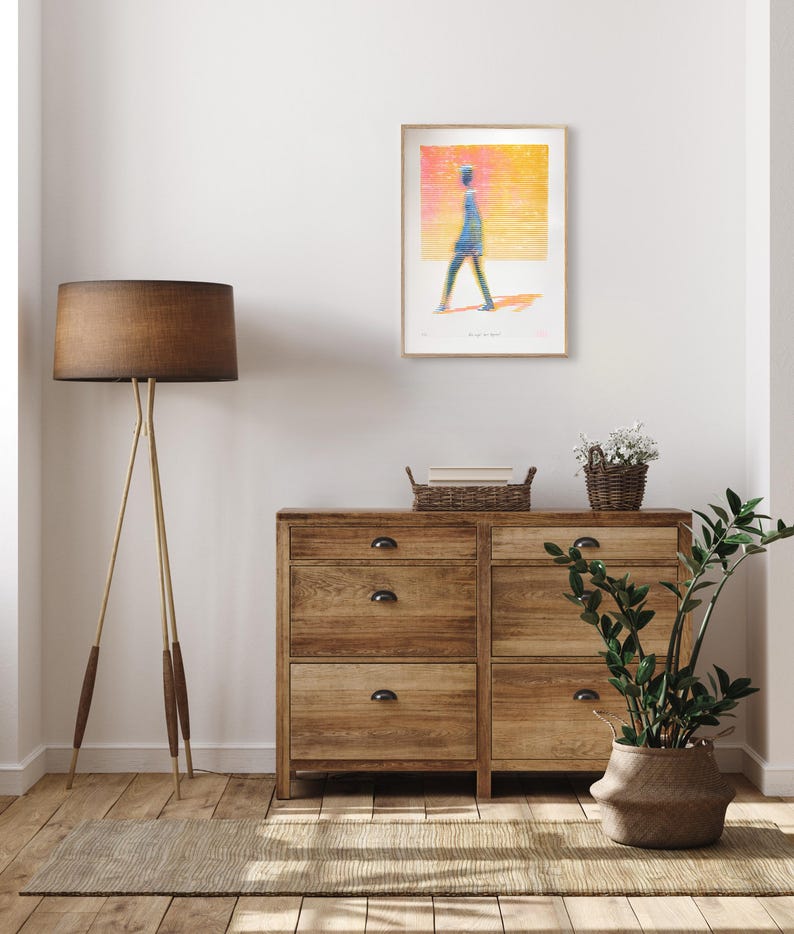 K&ouml;nnte beinhalten: Ein gerahmter Kunstdruck mit einer stilisierten Figur, die vor einem leuchtend gelben und orangen Hintergrund geht, an einer Wand &uuml;ber einer h&ouml;lzernen Kommode mit sechs Schubladen, einer Stehlampe, einem geflochtenen Korb und einer Pflanze.