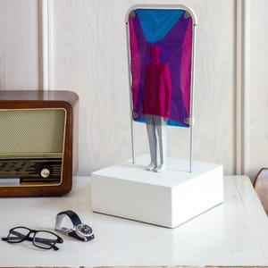Puede incluir: Una escultura que presenta una figura gris sobre una base rectangular blanca. Detr&aacute;s de la figura hay una estructura alta con marco blanco que sostiene una tela de malla transl&uacute;cida en tonos magenta, morado y azul brillante.