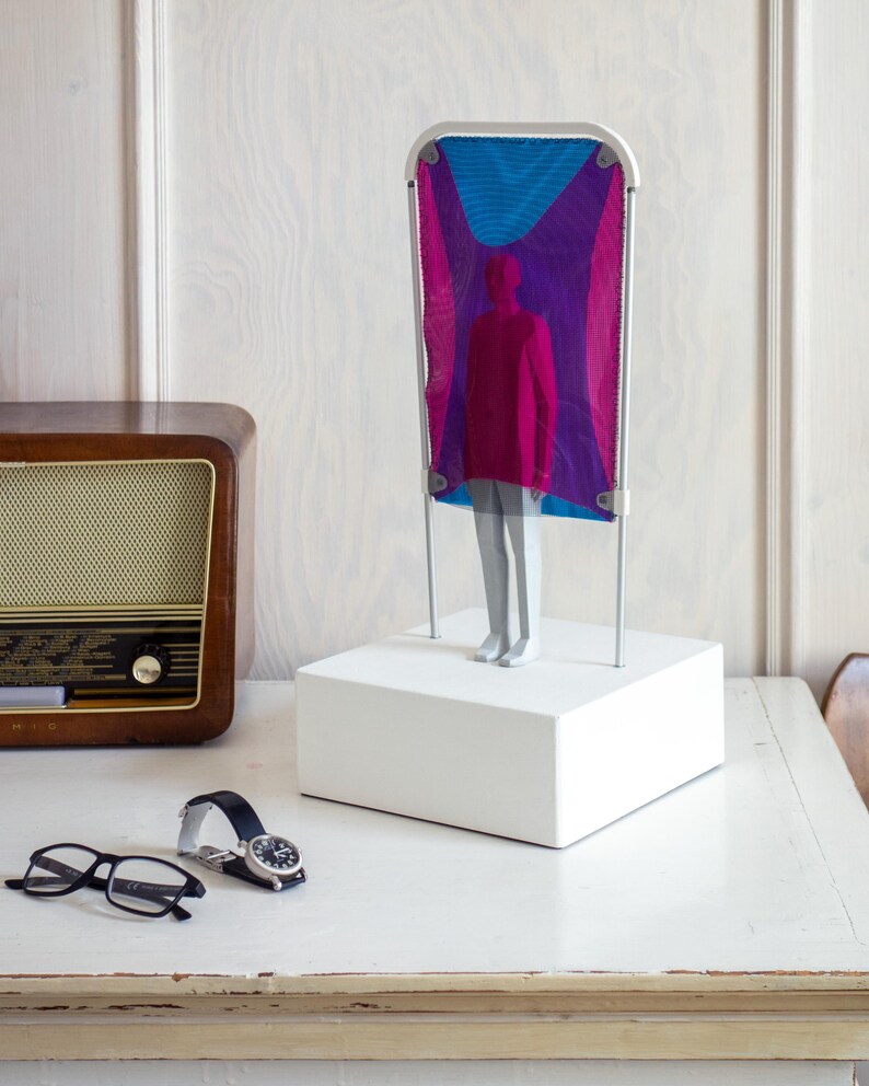 Puede incluir: Una escultura que presenta una figura gris sobre una base rectangular blanca. Detr&aacute;s de la figura hay una estructura alta con marco blanco que sostiene una tela de malla transl&uacute;cida en tonos magenta, morado y azul brillante.