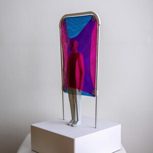 Puede incluir: Una escultura con una figura blanca de pie sobre una base rectangular blanca. Detr&aacute;s de la figura hay un marco de metal blanco que sostiene una malla transl&uacute;cida con degradados de colores azul, magenta y violeta sobre la figura.