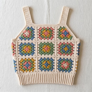 Puede incluir: Un top sin mangas de ganchillo construido a partir de múltiples motivos cuadrados con una base de color crema. Cada cuadrado contiene un patrón central de hilo rosa, amarillo, azul y verde. La prenda incluye tirantes anchos y un dobladillo acanalado.