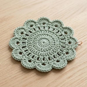 May include: A sage green, handmade crochet coaster with a scalloped edge. The coaster features a spiral design in the center and a small copper-colored ring attached to the edge. The coaster is placed on a light wood surface.