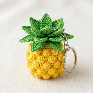 May include: A handmade crochet pineapple keychain. The pineapple is yellow with a textured surface, topped with green leaves. A silver-colored chain and keyring are attached. The keychain is a tropical-themed accessory.
