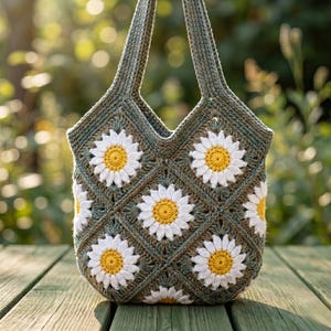 Könnte beinhalten: Eine handgefertigte Häkeltasche mit einem Gänseblümchenmuster. Die Tasche ist olivgrün mit weißen und gelben Gänseblümchen-Applikationen. Die Tasche hat zwei Henkel und steht auf einer Holzoberfläche.