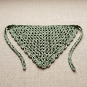 May include: A sage green crochet bandana with a triangular shape and scalloped edges. The open weave design features a diamond pattern. The bandana has long ties for securing around the neck or head.